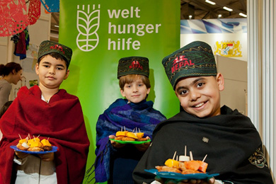 Das Foto ziegt drei Kinder in Trachten, sie haben Teller mit Speisen in den Händen. Im Hintergrund ist eine Fahne der Welthungerhilfe zu sehen. Die Szene ist auf der Bildungsmesse didacta aufgenommen.