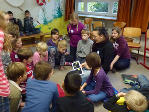 Das Foto zeigt Kinder, die auf dem Fußboden ihres Klassenraumes sitzen und denen eine Umweltpädagogin anhand eines Bilderbuches den Treibhauseffekt erklärt.