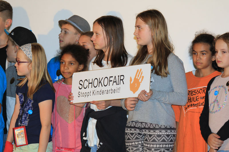 Das Foto zeigt eine Gruppe Kinder auf einer Bühne. Eines hat ein Schild mit der Aufschrift Schokofair-Projekt.