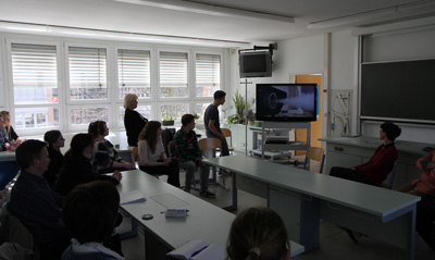 Das Foto zeigt Sch&uuler und erwachsene Personen in einem Klassenraum. Sie schauen auf einen Fernseher, darin ist eine Nebelmaschine zu sehen, welche Nebel ausstößt.