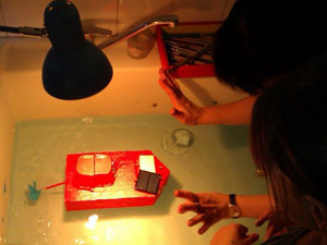 Das Foto zeigt den Blick in eine Badewanne, in dem ein kleines rotes Bootsmodell fährt. Es wird mit einem Elektromotor angetrieben, der seinen Strom von einer Solarzelle bezieht. Eine Lampe beleuchtet die Solarzelle. Zwei Kinder beugen sich über die Wanne und sehen zu.