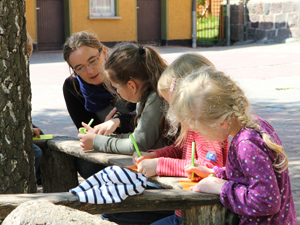 Das Foto zeigt drei Kinder, die auf einer Bank auf einem Schulhof konzentriert schreiben, sowie eine Frau, die ihnen zusieht.