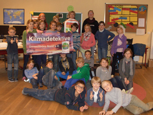 Das Foto zeigt eine Gruppe von Kindern, die in ihrem Klassenraum zum Gruppenbild posieren. Sie halten ein Transparent mit der Audschrift Klimadetektive.