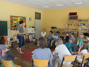 Das Foto zeigt eine Gruppe von 12 Personen - Kindern und Erwachsenen - die im Stuhlkreis in einem Klassenraum sitzen. Eine Frau steht.