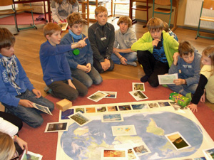 Das Foto zeigt eine Gruppe von Schüern, die auf dem Fußboden um eine Weltkarte herum sitzen. Sie halten Fotos in der Hand und ordnen diese auf der Weltkarte an.