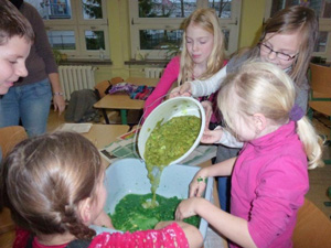 Das Foto zeigt 5 Kinder, die eine grünliche Pampe aus einer Schüssel in eine Wanne kippen.