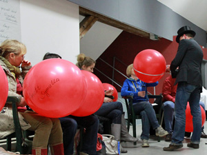 Das Foto zeigt einige Menschen, die auf Stühlen sitzen und große rote Luftballons in den Händen halten. Ein Mann mit Jackett und Zylinder misst den Umfang der Ballons.