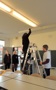 Das Bild zeigt einen Klassenraum in einer Schule. Eine Frau steht auf einer Leiter und greift zur Deckenlampe. Zwei Schüler halten die Leiter. Zwei Frauen schauen zu.