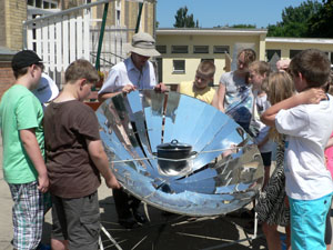 Das Foto zeigt Schüler, die im einen Solarkocher herum stehen. Ein Mann mit Sonnenhut erklärt ihnen den Kocher.