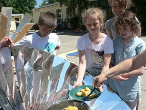 Das Foto zeigt Schüler, die an einen Solarkocher stehen und Maisscheiben aus dem Kochtopf nehmen.