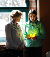 Das Foto zeigt zwei Frauen, die vor einem Fenster stehen und sich unterhalten. Eine hält einen gelben Zettel in der Hand, der von hinten von der Sonne zum Leuchten gebracht wird