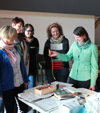 Das Foto zeigt fünf Frauen, die um einen Tisch herum stehen. Eine Frau hält ein Stromkabel in der Hand und erklärt den anderen etwas.