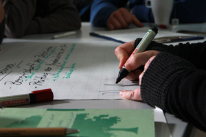 Das Foto zeigt einen Tisch. Darauf liegt Papier. Man sieht ferner die Hände von zwei Menschen, die mit dicken Stiften auf das Papier schreiben.