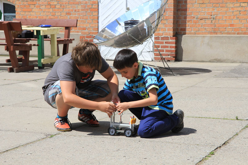 Das Foto zeigt zwei Schüler, die auf einem sonnigen Schulhof Solarfahrzeuge testen.