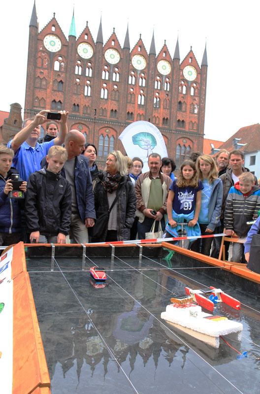 Das Foto zeigt Menschen, die um ein Wasserbecken herum stehen. Darin schwimmen kleine Solarboote. Im Hintergrund ist das Rathaus der Hansestadt Stralsund zu sehen.
