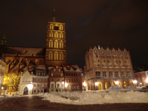 Das Foto zeigt den Alten Markt in Stralsund mit dem Rathaus und der Nikolaikirche, bei Nacht, festlich angestrahlt.