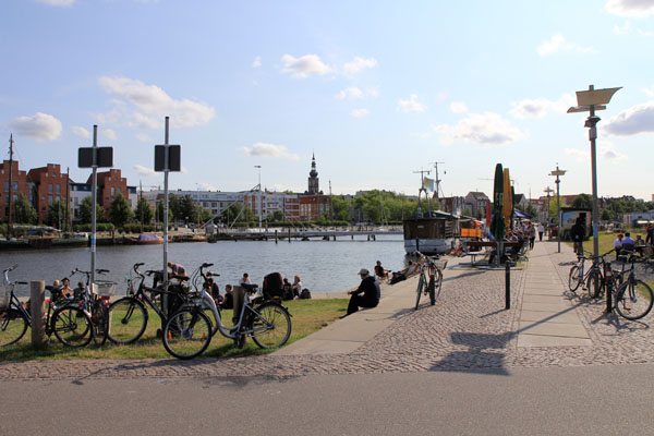 Das Foto zeigt eine Flusspromenade und einen Fluss - den Ryk in Greifswald - in einer sommerhellen Stimmung.