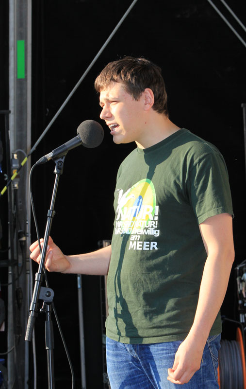 Das Foto zeigt einen Mann, der auf einer Bühne steht und in ein Mikrofon spricht.