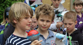 Das Foto zeigt einige Kinder auf dem Schulhof. Ein Junge hat ein Spielzeugmikrofon in der Hand und spricht in dieses hinein. Die anderen lauschen ihm; sie haben Bastelarbeiten - Windmühlen und Solarfinger - in den Händen.