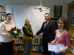 Das Foto zeigt drei Frauen und einen Mann bei der Übergabe eines Schecks. Im Hintergrund sieht man den Bibliotheksraum  mit Bücherregalen.