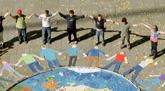Das Foto zeigt die Aktion Wir malen und Mensch und Erde aus mit dem Künstler Claus Pulm. Es sind Menschen zu sehen, die sich an den Händen halten und, einen großen Kreis bildend, um eine gemalte Erdkugel legensgroße menschliche Figuren herum stehen.